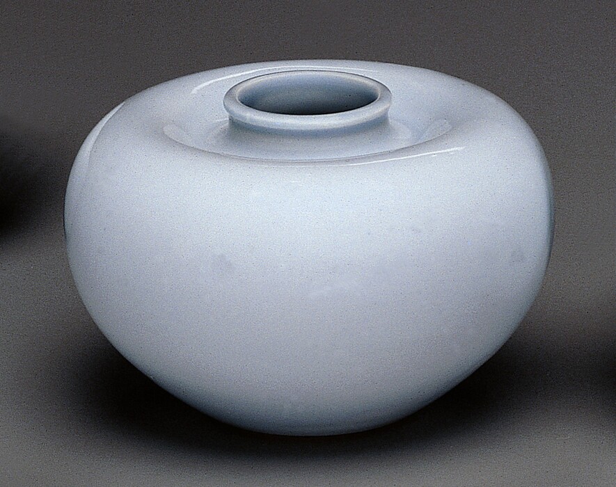 The sculpture is a globular-shaped ceramic jar with a narrow, slightly elevated neck. It has a smooth surface with an even, light blue color. The jar's shape is symmetrical, with a rounded lower body that tapers up to a smaller, circular opening. The material appears to be a glazed porcelain or similar ceramic, evident from its glossy finish that reflects light subtly across its surface. The overall look of the jar is simple and elegant, with a harmonious form that complements its monochromatic color.