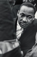 The image shows a man's upper body, focusing on his face and shoulders. He is leaning slightly forward, turning his head to his left, with his gaze directed towards the camera. The man has a distinct facial structure with a broad nose and full lips. He has short, closely cropped hair. He is wearing a formal suit jacket with a white dress shirt underneath and a patterned tie. Another person's head is partially visible in the foreground and there are indistinct figures in the background. The photograph is black and white, with the background out of focus.