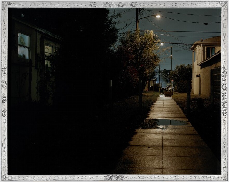 The image is captured from ground-level, showing a narrow alleyway in a residential area. The alley stretches into the distance with bends, bordered by buildings on one side and bushes and trees on the other. Streetlights illuminate the path, including one close by and a couple further away, casting a warm glow on the wet pavement. Puddles of water reflect the light. A dimly-lit house on the left has faint light from its windows, indicating nighttime. The scene features deep shadows and soft, warm artificial lighting, creating a muted yet contrasting night setting.