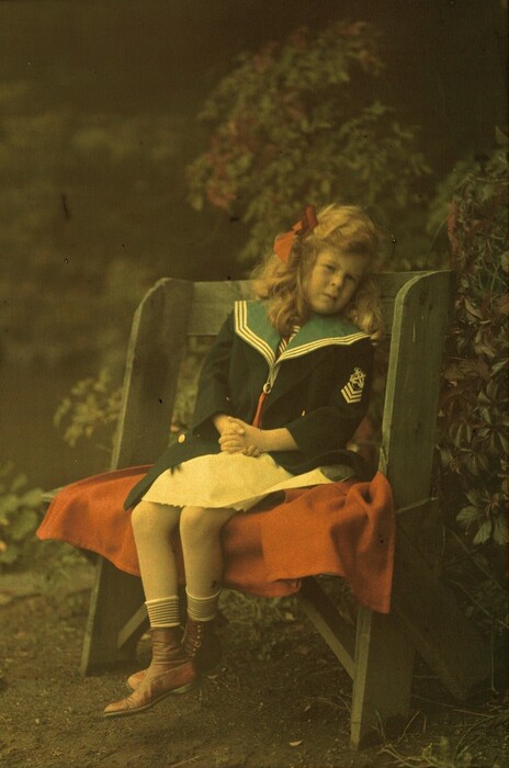 The image depicts a girl sitting on a wooden bench with a red blanket draped over it. She has long, curly blond hair with a red bow, and is wearing a navy blue sailor-style jacket with a light blue collar, a cream-colored skirt, knee-high striped socks, and brown boots. The background shows dense foliage and greenery around the base of the bench.