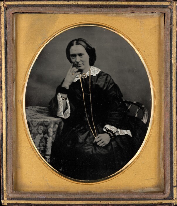 The image shows a woman seated with her upper body visible. She is posed with her elbow resting on a table draped with a patterned cloth, her hand touching her face, and adorned with jewelry on her fingers. Her facial features are distinct, with a broad forehead and soft expression, accentuated by her hair parted in the middle and styled smoothly to frame her face. She is dressed in a dark gown with a high neckline, white lace collar, cuffs of matching lace, and a long chain necklace. The background is plain, and the image is presented in an oval frame with a gold-colored border.