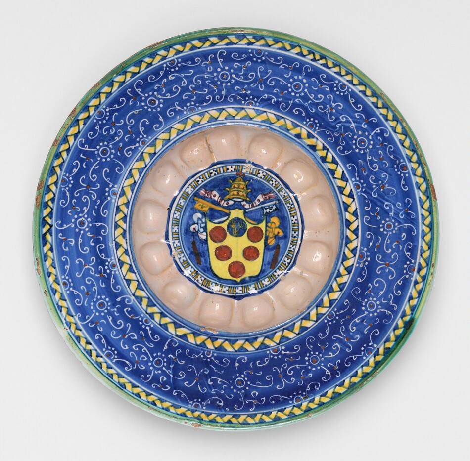 This is a photograph of a decorative ceramic plate with intricate designs. The plate features a central emblem with a shield design, displaying five red roundels on a golden field, topped by a papal tiara and crossed keys. Surrounding the emblem is an ornate pattern with a blue background and white swirling motifs. The outer border of the plate is adorned with alternating green and yellow geometric designs. The craftsmanship suggests a detailed and meticulous approach to decoration, likely intended for ornamental use.