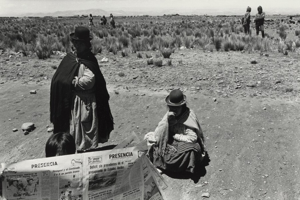 This is a black and white photograph of a rural landscape featuring people in traditional attire. The setting appears dry and arid with sparse vegetation. In the foreground, one person is holding a newspaper while sitting, and another is seated nearby wearing a dark hat and shawl. To the left and slightly further back, another individual stands in traditional clothing with a shawl and hat. The background shows a vast field with tufts of grass and small rocks, and in the distance, a few figures are visible standing or walking along the horizon.