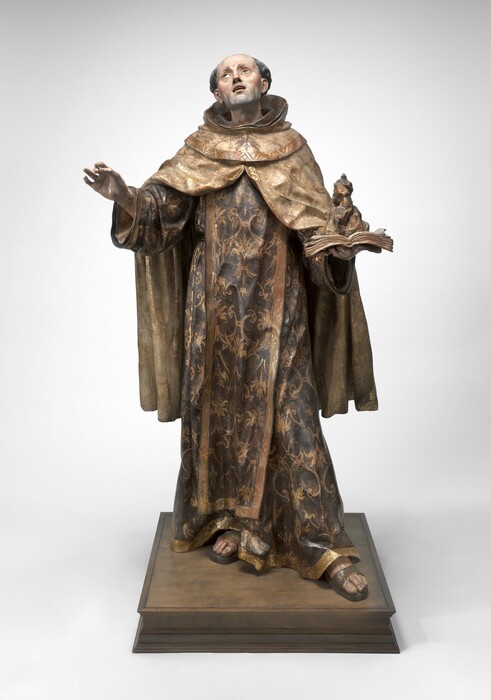 A pale-skinned man wearing voluminous robes under a monk’s hood is carved from wood and painted in this freestanding sculpture. In this photograph, the man’s body faces us as he tips his head to our left and back to look up in that direction. His lips are parted, and his square chin is shaded with a five o’clock shadow. His gray hair is curly over each ear, and the top of his head is bald. He holds his right hand, to our left, up with his thumb and first two fingers almost touching. He holds an open book in the flat of his other hand. A rocky formation on the pages, the edges of which curl up, could be a flame. The man’s hood is pushed back, and the ivory-white, knee-length cloak drapes heavily over his shoulders and parts over his spread arms. He wears a long robe painted earthy brown that reaches his sandaled feet. Both the cloak and robe are edged with gold along the hems and painted with gold, leafy designs. The sleeves of his robe hang wide around his wrists. He stands with his weight on his right foot and the other foot thrust a bit forward on a square, wood base. The sculpture is photographed against a white background.