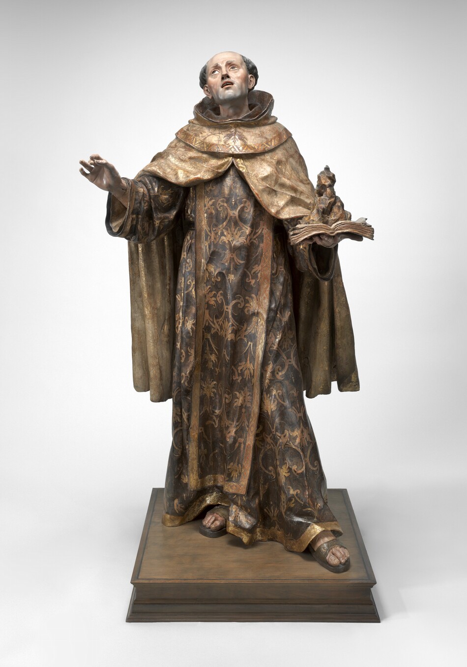 A pale-skinned man wearing voluminous robes under a monk’s hood is carved from wood and painted in this freestanding sculpture. In this photograph, the man’s body faces us as he tips his head to our left and back to look up in that direction. His lips are parted, and his square chin is shaded with a five o’clock shadow. His gray hair is curly over each ear, and the top of his head is bald. He holds his right hand, to our left, up with his thumb and first two fingers almost touching. He holds an open book in the flat of his other hand. A rocky formation on the pages, the edges of which curl up, could be a flame. The man’s hood is pushed back, and the ivory-white, knee-length cloak drapes heavily over his shoulders and parts over his spread arms. He wears a long robe painted earthy brown that reaches his sandaled feet. Both the cloak and robe are edged with gold along the hems and painted with gold, leafy designs. The sleeves of his robe hang wide around his wrists. He stands with his weight on his right foot and the other foot thrust a bit forward on a square, wood base. The sculpture is photographed against a white background.