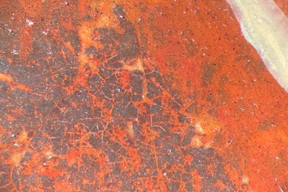 A photomicrograph view of blackened vermilion, showing an overall field of predominant reddish orange with dark grey splotches in a diagonal strip, speckled with lighter white spots and a broad white strip in the top right corner