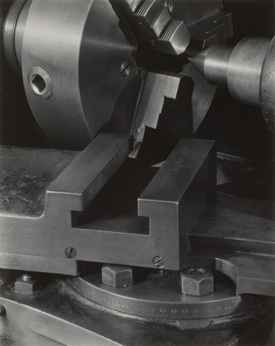 This is a photograph of a metal lathe chuck and vise. The image showcases a close-up perspective of the machinery setup, including the chuck, jaws, and a vise. The metal components have precise edges and industrial screws. The metal surface is reflective, highlighting the polished steel. The photograph likely focuses on the intricacy and precision involved in mechanical instrument design.