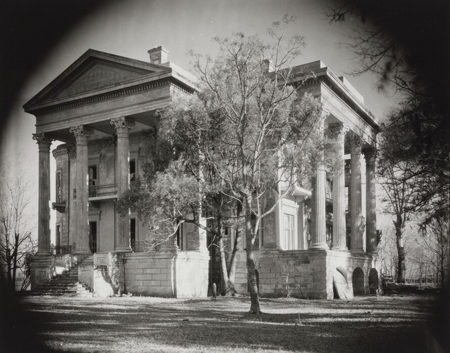 The image shows a grand neoclassical mansion viewed slightly from the side. The stone staircase, tall Corinthian columns, and detailed pediment adorn the entrance, which is elevated and surrounded by railings. The horizon is not distinctly visible. The mansion is partially obscured by tall, leafless trees that cast shadows. The photograph is in black-and-white, emphasizing the texture of the stone, shadows, and architecture. The sky appears clear in the background.