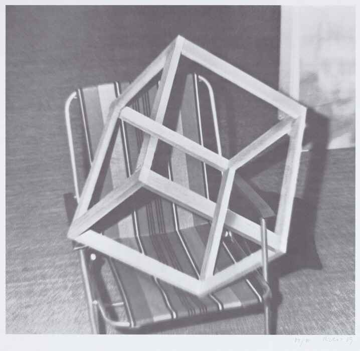 The image features a complex geometric shape made up of interlocking rectangular frames placed on a striped folding chair. The arrangement creates an abstract design, with lines and shadows adding depth to the structure.