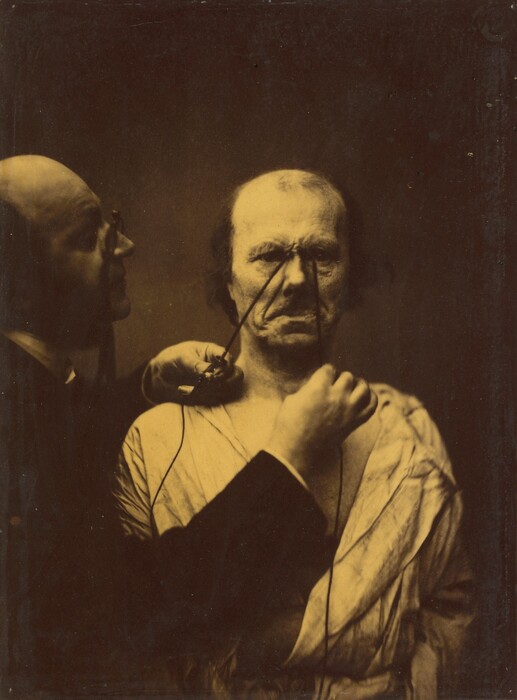 This is a photograph of a man conducting an experiment with electrical apparatus on a model or mannequin. The photograph features a bald figure on the left adjusting something connected to another mannequin or model in the center. Thin wires are attached to the central figure's face, leading back towards the hand of the left figure.