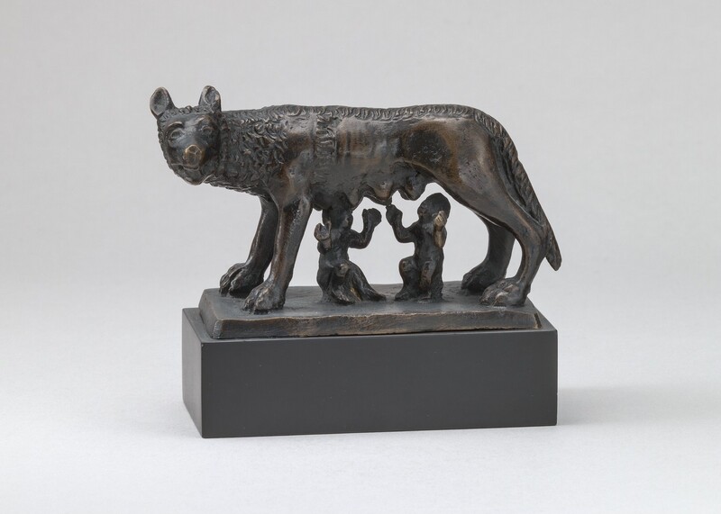 A freestanding, bronze she-wolf stands as two male infants attempt to nurse at her teats in this sculpture. In this photograph, the wolf stands sideways facing our left with her head turned to look at us, eight teats hanging heavily along her underside. Her eyes and brows are represented with ridges but her features are worn. Her jaws are slightly open, and her lean body is partly covered by tight clusters curls around her head, neck, and along her back. Beneath her, the boys are roughly modeled or perhaps also worn. They both appear to sit on rocks as they tilt their heads back and spread their arms wide. The boys and wolf are on a slab of bronze, which in turn rests on a gray, rectangular base. The sculpture is photographed in front of a white wall.