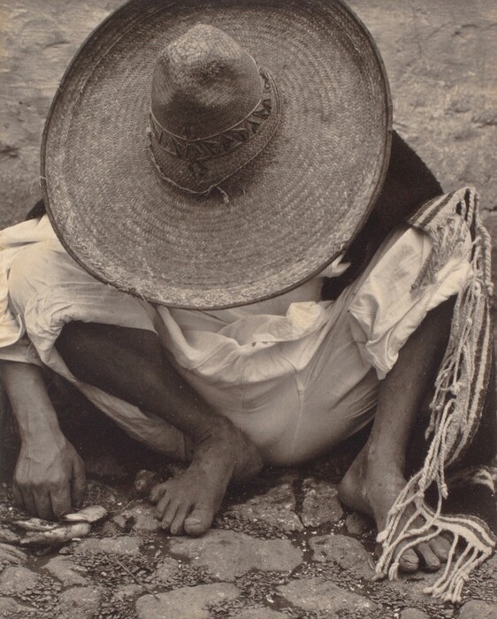 The image shows a person seated on a cobblestone path, wearing a sombrero that covers their head and face. They are dressed in a loose shirt, pants, and a woven cloth draped around their body. The person's head is bent down, hiding their face while they are holding small objects in their left hand.