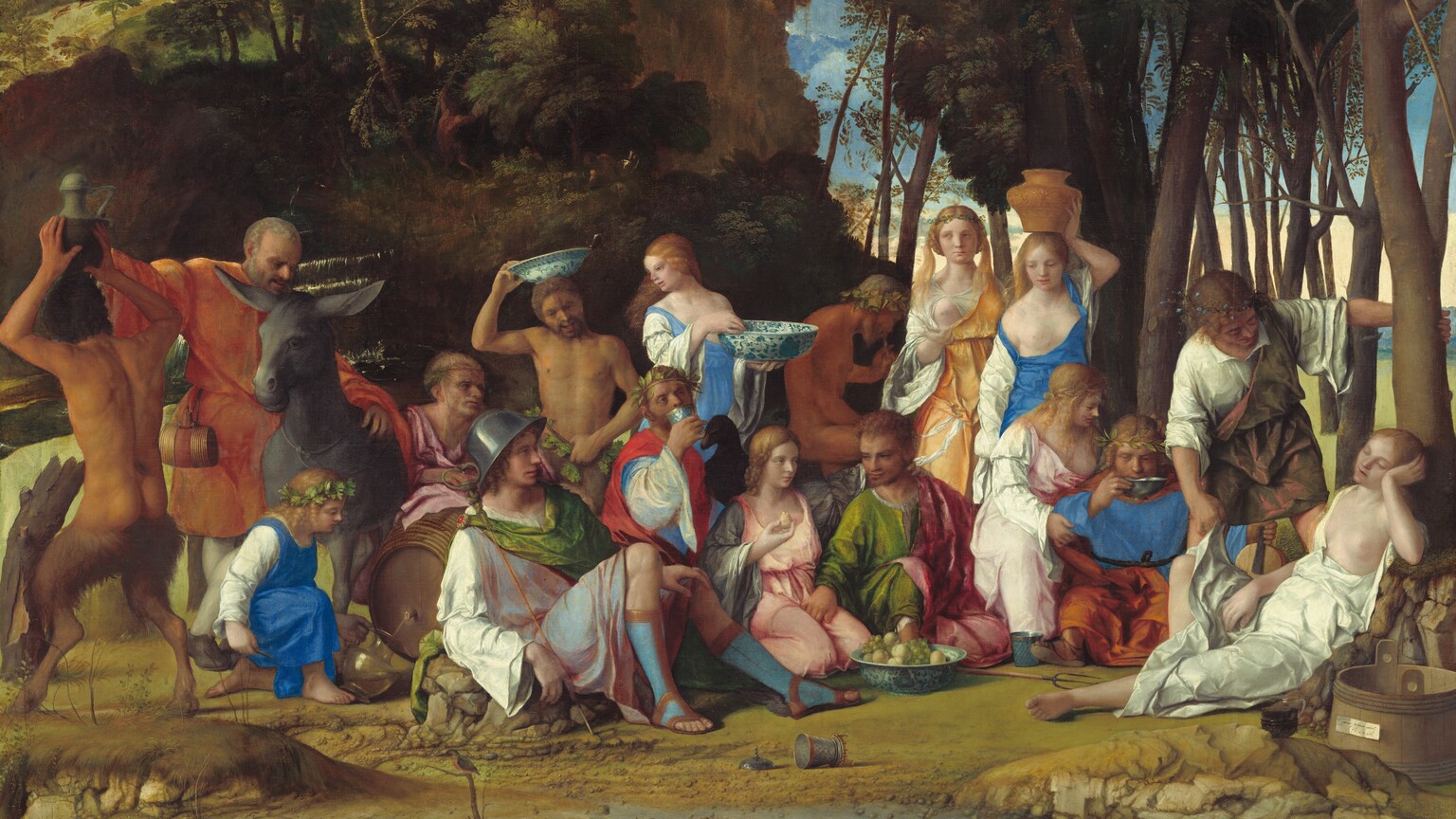 Six women, eight men, two satyrs, and one child gather in pairs and trios in a loose row that spans the width of this nearly square painting. They are set within a landscape with craggy rocks, cliffs, and trees. Most of the people face us, and the men, women, and child have pale skin. The two satyrs have men’s torsos and furry goat’s legs, and they have darker, olive complexions. Most of the men wear voluminous, knee-length togas wrapped in short robes in shades of white, topaz blue, grass green, coral orange, or rose pink. Most of the women wear long, dress-like garments in tones of shell pink, apricot orange, or lapis blue over white sleeves. For all but one woman, their garments have fallen off one shoulder to reveal a round, firm breast. Several objects are strewn on the rocky, dirt ground in front of the group, including a wide, wooden bucket with a piece of paper affixed to its front to our right, a glass goblet, a pitchfork, a large blue and white ceramic dish filled with grapes and small yellow fruits, and an overturned cup near the center. Cliff-like, craggy rocks rise steeply behind the group to our left, filling much of the sky opposite a tall grove of leafy, dark green trees to our right. A few puffy white clouds float across the vivid blue sky. The slip of paper on the barrel has been inscribed, “joannes bellinus venetus p MDXIIII.”