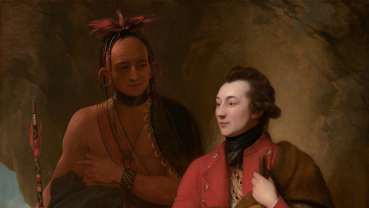 A man with pale white skin sits and a man with light brown skin stands in front of a cave-like, rocky outcropping in this vertical portrait painting. The light-skinned man closer to us has dark brown hair, and the light falls more strongly on him. He sits propped on or against a rock with his body angled to our left, and he looks off into the distance in that direction. He wears a scarlet-red overcoat, a white waistcoat, and moccasins. What appears to be an animal skin painted on the underside with red, tan, and black geometric patterns is tied around his chest and falls over his left arm, on our right. With that hand, he holds a musket like a walking stick. He also holds a black cap ornamented with beads and feathers on his other knee. The man with brown skin stands in shadow behind the other man, to our left. His body faces us, and he looks at the seated man. A dark cloak encircles his shoulders and is gathered around his waist. A strap crosses his bare chest and the strap, bracelets, and a feathered headdress seem to be beaded. Large gold rings curve from his earlobes up over his ears. A puff of smoke emerges from the top of the long, painted stick he holds, suggesting it is a pipe. He holds his left hand, on our right, at his chest, and points subtly to our left, perhaps to the landscape seen through a break in the rocky outcropping. There, in front of a waterfall in the distance, a small group of people with light brown skin gather around a camp fire in front of a tent-like structure.