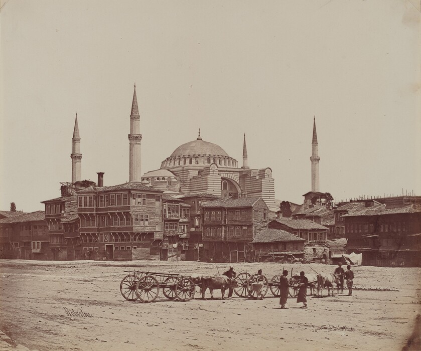The image shows a cityscape looking towards a busy street. People guide carts pulled by oxen on a dirt path in the foreground. In the center, a mosque with a large dome and four minarets stands out. The skyline is above the buildings, with wooden structures lining the street. The scene is in sepia tones, emphasizing shades of brown.
