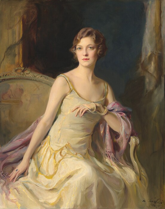 Shown from the knees up, a pale-skinned woman sits and leans on the arm of a seat in this vertical portrait painting. The woman’s knees and shoulders are angled to our right but she turns her head slightly to our left so she looks out at us from the corners of her green, wide-set eyes. She has an oval face with a short nose, full pink lips, and a cleft in her chin. Her brown hair is set in loose finger waves and is either chin-length or pulled back. Her sleeveless, scoop-necked, ivory-white gown has spaghetti straps edged with gold. A sheen on the fabric suggests that it shimmers between white, mauve pink, and gold, and gold tracery around her hips mimics the look of a drop-waist over a long, flaring skirt. A rose-pink shawl drapes over her left arm, to our right, which rests on the arm of her seat. A long strand of pearls is looped twice around that wrist like a bracelet as the other end is pulled over her fingers, which she holds in front of her chest. Her other arm, to our left, is stretched straight by her side, and she wears a ring on that hand. The seat has an arched back and is painted with streaks of gold and tan. A darkened opening behind her is shaded midnight blue. Gold frames or molding flank the opening along the left and right edges of the canvas. The portrait is loosely painted, especially in her clothing and the background. The artist signed and dated the bottom right corner, “de Lászlo 1626. III.”