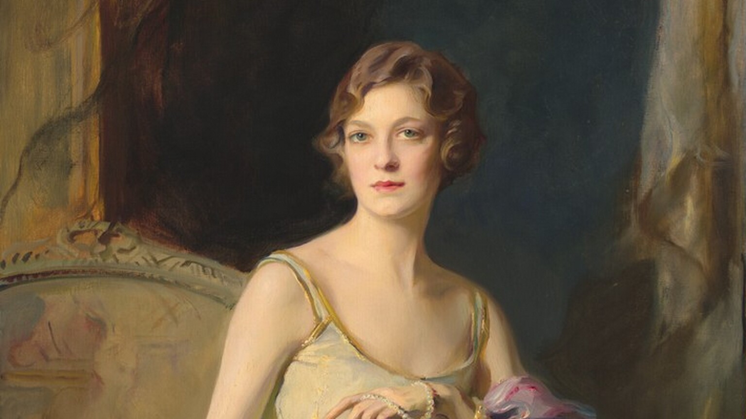 Shown from the knees up, a pale-skinned woman sits and leans on the arm of a seat in this vertical portrait painting. The woman’s knees and shoulders are angled to our right but she turns her head slightly to our left so she looks out at us from the corners of her green, wide-set eyes. She has an oval face with a short nose, full pink lips, and a cleft in her chin. Her brown hair is set in loose finger waves and is either chin-length or pulled back. Her sleeveless, scoop-necked, ivory-white gown has spaghetti straps edged with gold. A sheen on the fabric suggests that it shimmers between white, mauve pink, and gold, and gold tracery around her hips mimics the look of a drop-waist over a long, flaring skirt. A rose-pink shawl drapes over her left arm, to our right, which rests on the arm of her seat. A long strand of pearls is looped twice around that wrist like a bracelet as the other end is pulled over her fingers, which she holds in front of her chest. Her other arm, to our left, is stretched straight by her side, and she wears a ring on that hand. The seat has an arched back and is painted with streaks of gold and tan. A darkened opening behind her is shaded midnight blue. Gold frames or molding flank the opening along the left and right edges of the canvas. The portrait is loosely painted, especially in her clothing and the background. The artist signed and dated the bottom right corner, “de Lászlo 1626. III.”