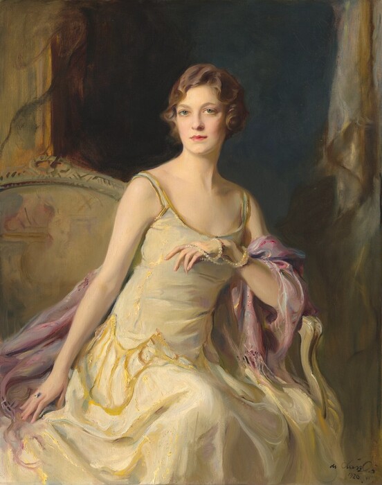 Shown from the knees up, a pale-skinned woman sits and leans on the arm of a seat in this vertical portrait painting. The woman’s knees and shoulders are angled to our right but she turns her head slightly to our left so she looks out at us from the corners of her green, wide-set eyes. She has an oval face with a short nose, full pink lips, and a cleft in her chin. Her brown hair is set in loose finger waves and is either chin-length or pulled back. Her sleeveless, scoop-necked, ivory-white gown has spaghetti straps edged with gold. A sheen on the fabric suggests that it shimmers between white, mauve pink, and gold, and gold tracery around her hips mimics the look of a drop-waist over a long, flaring skirt. A rose-pink shawl drapes over her left arm, to our right, which rests on the arm of her seat. A long strand of pearls is looped twice around that wrist like a bracelet as the other end is pulled over her fingers, which she holds in front of her chest. Her other arm, to our left, is stretched straight by her side, and she wears a ring on that hand. The seat has an arched back and is painted with streaks of gold and tan. A darkened opening behind her is shaded midnight blue. Gold frames or molding flank the opening along the left and right edges of the canvas. The portrait is loosely painted, especially in her clothing and the background. The artist signed and dated the bottom right corner, “de Lászlo 1626. III.”