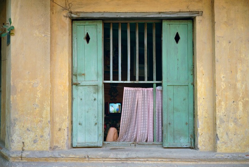 In this photograph, a weathered, vertical rectangular window is framed by a yellowish, textured wall that exhibits signs of aging and wear. The window features two hinged green shutters, each with a diamond-shaped cutout near the top. Behind the shutters, metal bars span the top of the window opening. Below, a pink and white checkered curtain partially blocks the view. On the left, a bent leg is visible, resting beside the curtain, as if someone is sitting inside with their back against the shutter. Further inside, a television screen displays an image that is hard to make out.