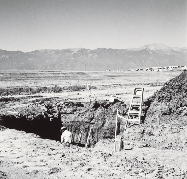 The image shows a black and white landscape with a man working in a trench. The viewpoint is slightly elevated, revealing the vast terrain stretching towards the horizon. Mountains dominate the skyline in the distance. The landscape is desolate apart from the man working in the trench. A ladder leans against the trench wall. The photo emphasizes clarity and detail, showing textures of the dirt and subtle shadows. The mountains in the background add depth. The grayscale tones create a stark atmosphere, emphasizing the rugged terrain.