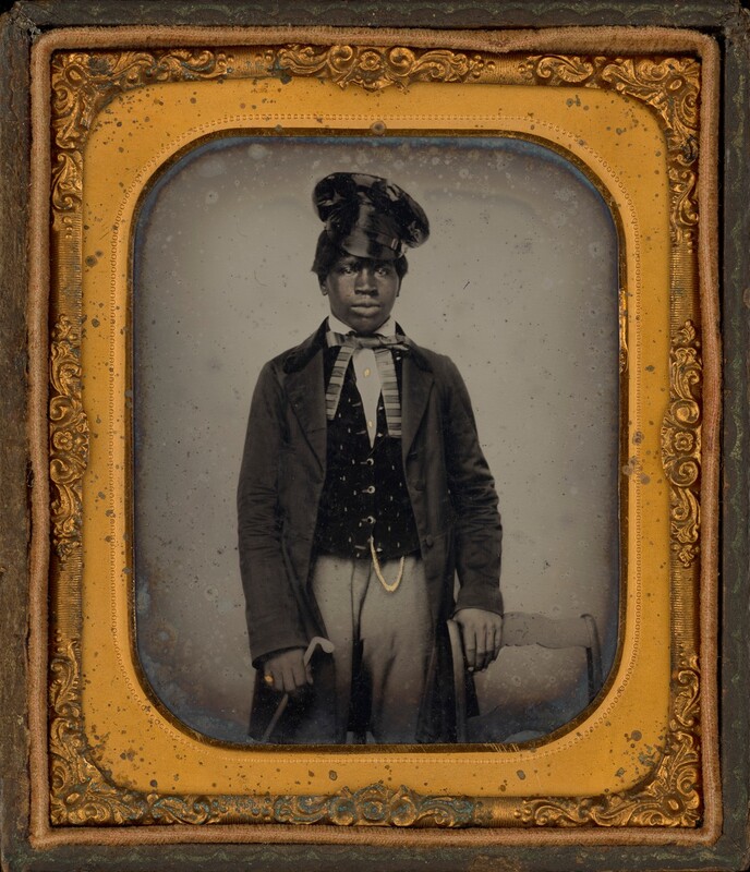 The image depicts a young person's upper body, from waist up, standing and slightly turned to their right while looking directly at the camera. They have a serious expression, rounded cheeks, full lips, and dark, short hair mostly hidden beneath a shiny top hat. They are dressed in a formal outfit consisting of a dark jacket, a patterned waistcoat with light-colored buttons, a white collared shirt, and a striped cravat. A watch chain is visible hanging from the waistcoat. They are holding a cane in their left hand and resting their right hand on the top of a chair beside them. The image is framed with an ornate golden frame with detailed decorations, enclosing the photograph. The background is a plain, light-colored surface.