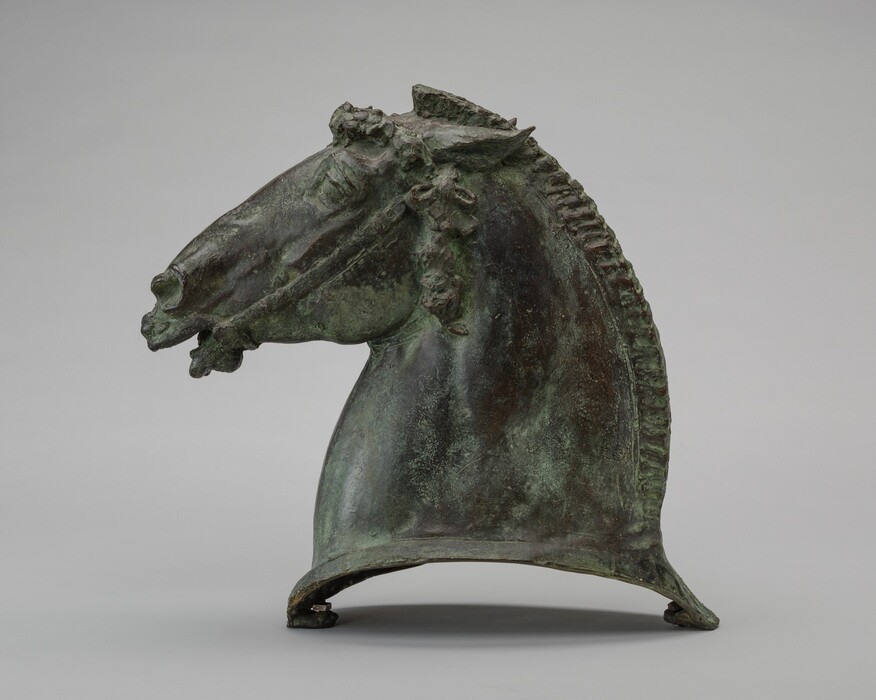The sculpture depicts a horse's head with intricate details like ears, eyes, and a stylized mane. It is in bronze with a greenish-brown patina. The horse's head is slightly abstract and stylized, depicting strength and elegance. The sculpture has an arched base. The overall aesthetic is ancient or classical, exuding an imposing and dignified presence.