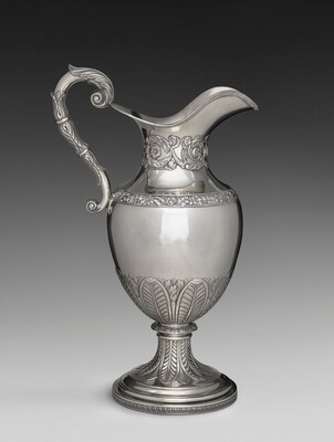The sculpture is of a jug or pitcher made of a metallic material, likely silver. It has a curved handle embellished with leaf and scroll motifs. The body is polished, with a decorative band near the neck featuring floral designs and an ornamental base with foliage patterns. The jug is tall, standing on a sturdy, decorated pedestal base. The metallic sculpture is sophisticated and luxurious, appearing functional and artistic.