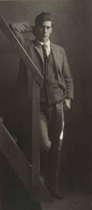 In the image, there is a man standing next to a wooden staircase. He is dressed in a three-piece suit with a white shirt and black tie. The man has a relaxed posture, with one arm on the staircase railing and the other on his hip. He has a strong jawline, distinct cheekbones, and dark, styled hair. The background is plain and dim, emphasizing the man.