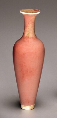 The sculpture is a vase with a long, slender neck and a rounded body that tapers towards the base. The vase is predominantly rose pink in color, with some slight variation on the neck, where there are patches of a peachy shade. There are also small dots of a light greenish color. The rim and base are both white.