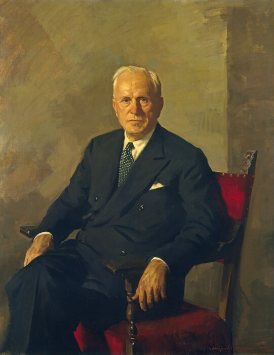 Shown from the shins up, a pale-skinned, cleanshaven man wearing a black suit sits and looks out at us in this vertical portrait painting. His legs are crossed and knees angled to our left, but he turns his face to us. He has a round face with slight bags under his gray eyes, and his thin lips are closed. His cheeks and jowls are a bit fleshy with age, and short white hair is brushed back from his high forehead. He wears a double-breasted jacket over a white shirt and dark blue tie with white polka dots. A white pocket square peeks out of the breast pocket. Both of the man’s elbows rest along the wood arms of his chair, which is upholstered on the back and seat with ruby-red fabric. The supports under the arms are turned, and top of the vertical style holding up the backrest that we can see is carved. The chair is near the corner of a room with walls painted muted sage green and tan. Brushstrokes are visible throughout and are especially loose in the background. The artist signed and dated the canvas with red paint in the lower right corner, “Leopold Seyffert 53.”