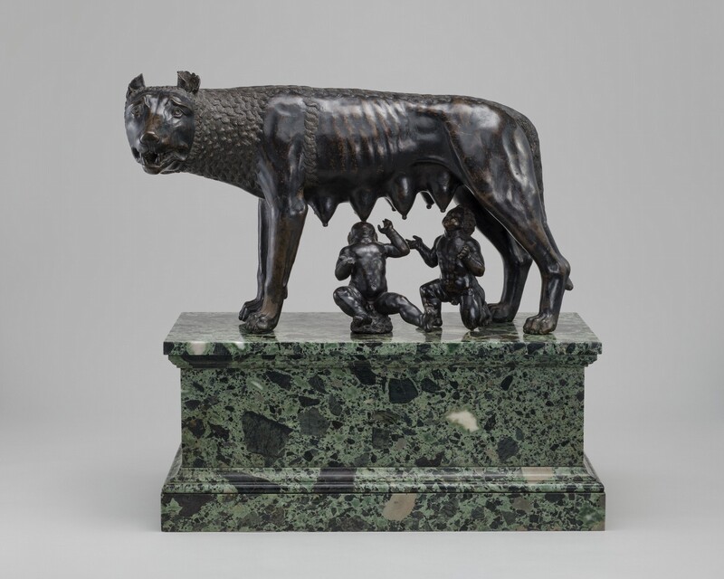 A free-standing, bronze she-wolf stands as two male infants attempt to nurse at her teats in this sculpture. In this photograph, the wolf stands sideways facing our left with her head turned to look at us, eight teats hanging heavily along her underside. Her wide eyes are framed by raised but furrowed brows, and cup-like ears curl back on her round head. Her jaws are slightly open revealing sharp teeth. Her lean body is partly covered by tight clusters of parallel, undulating lines around her head, neck, and along her back, suggesting fur. Beneath her, one boy sits, rocking back on a low rock, and the other kneels. Both tilt their heads back and spread their arms wide as they reach for her teats. The bronze of the boys and wolf are rubbed gold in some areas, especially on the wolf’s ribs. The trio rest on a green and black marble pedestal.