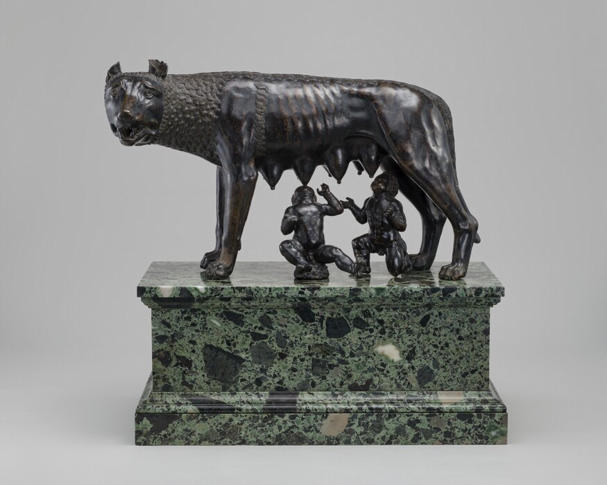 A free-standing, bronze she-wolf stands as two male infants attempt to nurse at her teats in this sculpture. In this photograph, the wolf stands sideways facing our left with her head turned to look at us, eight teats hanging heavily along her underside. Her wide eyes are framed by raised but furrowed brows, and cup-like ears curl back on her round head. Her jaws are slightly open revealing sharp teeth. Her lean body is partly covered by tight clusters of parallel, undulating lines around her head, neck, and along her back, suggesting fur. Beneath her, one boy sits, rocking back on a low rock, and the other kneels. Both tilt their heads back and spread their arms wide as they reach for her teats. The bronze of the boys and wolf are rubbed gold in some areas, especially on the wolf’s ribs. The trio rest on a green and black marble pedestal.