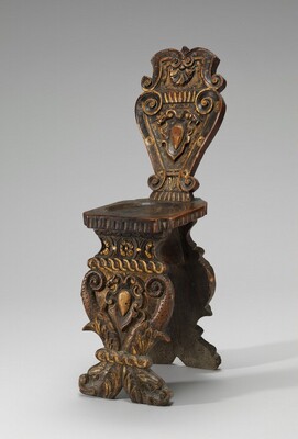 This is a photograph of an ornate wooden chair. The chair has intricate carvings and design elements throughout its structure. The backrest is detailed with scrollwork and central shields or cartouches framed by foliage designs. The seat is simple and flat, resting upon a similarly detailed base that continues the decorative motifs of scrolls and acanthus leaves. The chair appears to be made of dark wood, possibly enhanced with gold or gilded accents. The craftsmanship suggests it is an ornamental piece, possibly intended for decorative purposes as much as for utility.