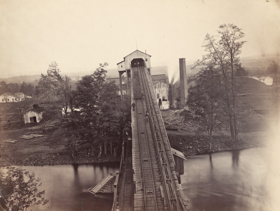 The image shows a wooden structure resembling an incline railway or conveyor leading towards a large building complex in the distance. A light haze obscures the horizon, hinting at an expansive forested area and other buildings. Trees on the sides blur softly, suggesting movement. A river with reflective waters flows close by. The setting appears historical and industrial, with a monochromatic color palette in shades of sepia and grey, giving it a vintage feel.