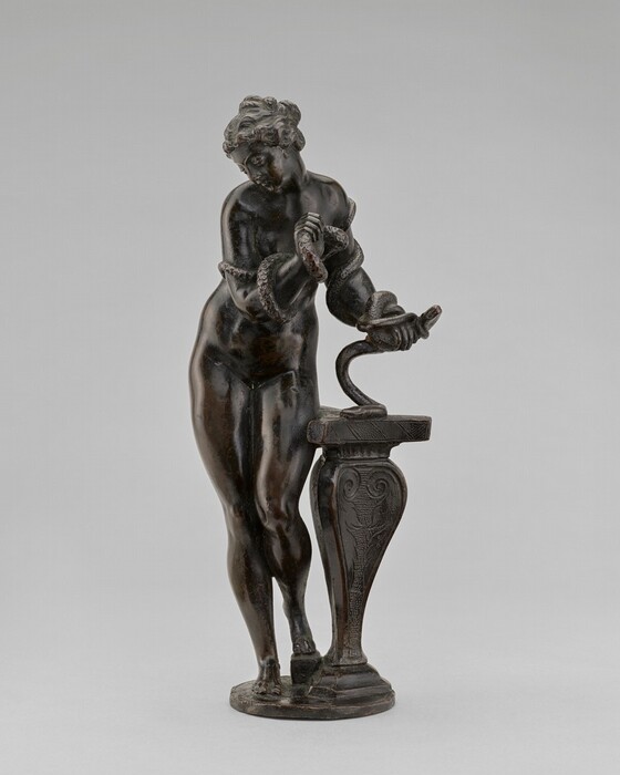 The sculpture depicts a standing nude figure of a woman with a robust, curvy body and curly hair. She is engaged in a dynamic pose, with her arms wrapped around a snake coiling around her body. The woman leans against a tall, intricately decorated pedestal, which features scrollwork and a sturdy base. The sculpture is crafted from a dark bronze material, which gives it a rich and lustrous appearance with subtle highlights and shadows. The figure's expression, along with her stance and the snake, creates a sense of movement and interaction within the composition. The sculpture is likely small to medium-sized, suitable for display on a mantel or within a collection of similar works.