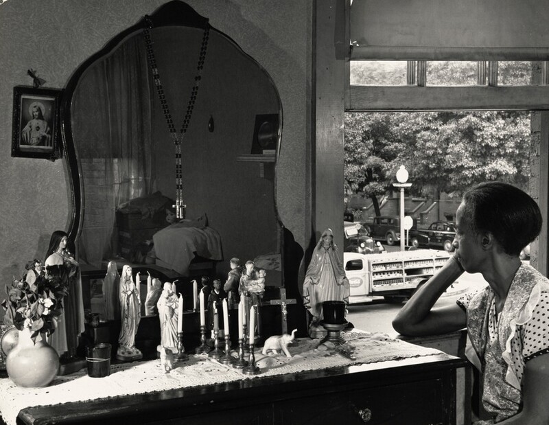 A Black woman sits facing a round-topped mirror on a dresser, which is crowded with religious figurines and candles in this horizontal black and white photograph. In the lower right corner, the woman rests her face in one hand, that elbow laid along the window frame of an open window. She wears a ruffle-edged apron over a short-sleeved garment with dark polka dots against white. Her face is turned almost away, and her hair is pulled back. Her eyes are lowered as she gazes at a figurine of the Virgin Mary on the dresser, which is covered with a lace runner. Starting from the left on the dresser are a gourd-shaped vase with dried flowers, two vessels, three figurines of saints, and a palm-sized statuette of an elephant before a row of five candles that separates the left half of the dresser-top from the right. Two more candles flank the row, and across the right half are a second elephant, two more religious figurines, two crucifixes, and a votive candle in front of the statuette of Mary. A rosary with a pendant-sized cross hangs from the curved top of the mirror, and a framed picture of Jesus hangs to the left. The mirror reflects part of the room behind the woman, showing shoeboxes, pieces of cloth, and a pale-faced doll stacked in the far corner of the small room. The window takes up the right half of the composition, and shows a view out to a street lined with trees, cars, a Coca-Cola delivery truck packed with glass bottles, a stop sign, and a streetlight.