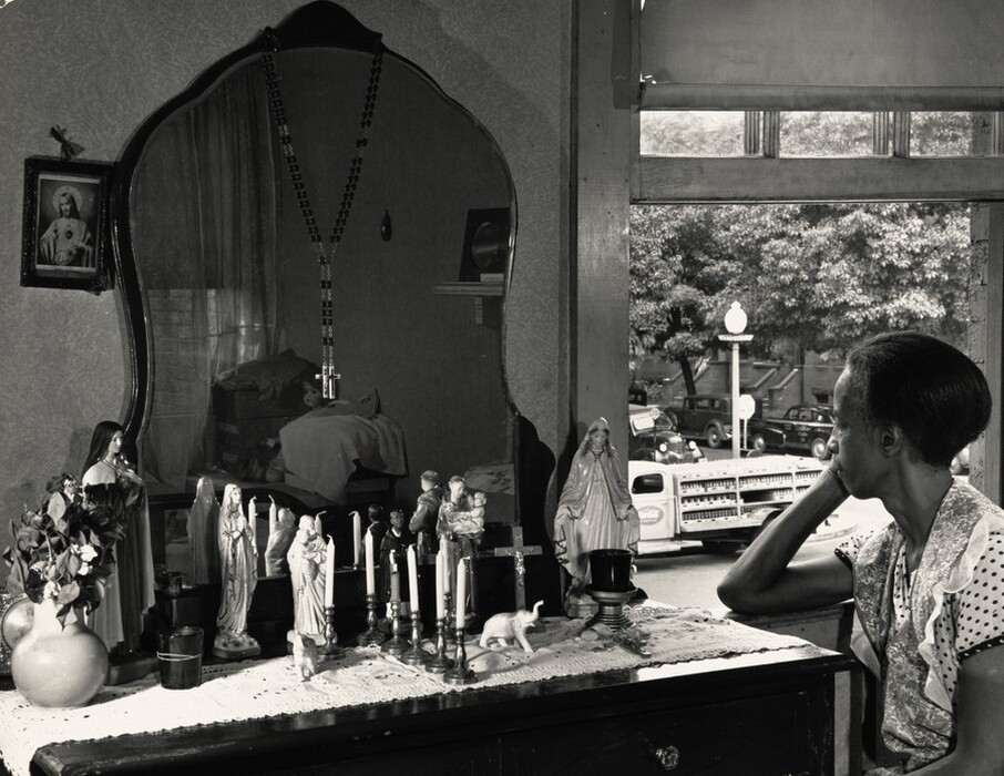 A Black woman sits facing a round-topped mirror on a dresser, which is crowded with religious figurines and candles in this horizontal black and white photograph. In the lower right corner, the woman rests her face in one hand, that elbow laid along the window frame of an open window. She wears a ruffle-edged apron over a short-sleeved garment with dark polka dots against white. Her face is turned almost away, and her hair is pulled back. Her eyes are lowered as she gazes at a figurine of the Virgin Mary on the dresser, which is covered with a lace runner. Starting from the left on the dresser are a gourd-shaped vase with dried flowers, two vessels, three figurines of saints, and a palm-sized statuette of an elephant before a row of five candles that separates the left half of the dresser-top from the right. Two more candles flank the row, and across the right half are a second elephant, two more religious figurines, two crucifixes, and a votive candle in front of the statuette of Mary. A rosary with a pendant-sized cross hangs from the curved top of the mirror, and a framed picture of Jesus hangs to the left. The mirror reflects part of the room behind the woman, showing shoeboxes, pieces of cloth, and a pale-faced doll stacked in the far corner of the small room. The window takes up the right half of the composition, and shows a view out to a street lined with trees, cars, a Coca-Cola delivery truck packed with glass bottles, a stop sign, and a streetlight.