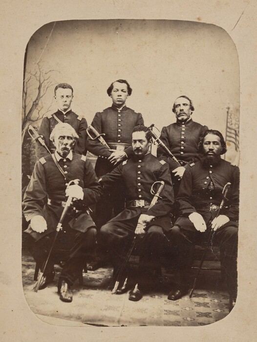 Six men in dark military uniforms are depicted in the image. The front row shows three seated men holding swords. The back row displays three standing men with serious expressions. The men have varying facial hair and hairstyles. They are all dressed in dark military uniforms with white collars and shoulder epaulets, holding swords. Some wear belts, indicating military rank. The background shows a painted backdrop with trees and an American flag. The setting appears to be indoors based on the patterned rug.