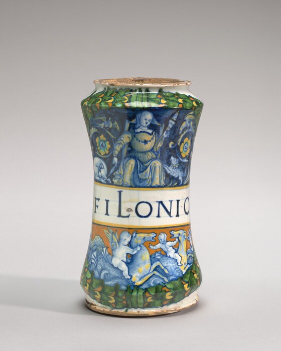 This is a photograph of a decorative ceramic vase. The vase has painted scenes and motifs across its surface. The upper portion features a seated figure, peacock-like designs, foliage, and an orange color amidst green leaves. The middle section has an inscription reading "FILIONIO." The bottom section depicts cherubs or putti, one riding on a dolphin-like creature over waves. The composition suggests an ornate, historical style, possibly reminiscent of Italian Renaissance ceramics known for vivid colors and detailed storytelling in art.