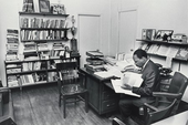 This is an image of an office space with a large bookshelf filled with books, trophies, a wooden desk with paperwork and books, and various decorations on the walls.