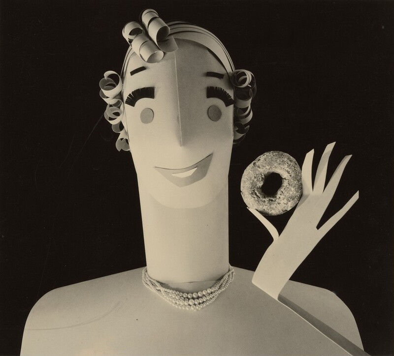 This is a photograph of a stylized paper sculpture depicting a figure. The image shows a paper sculpture figure with an abstract face, rolled paper for curls, a smile, and a forked hand holding a donut. The background is black, enhancing the figure's grayscale presentation.