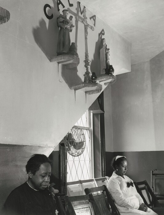 The image shows two seated women. One woman is in the foreground and is slightly to the left, while the other woman is in the background closer to a window. The woman in the foreground has her head slightly bowed with her eyes closed. She is wearing a dark-colored robe with a clerical collar visible and a rose pinned to her garment. The woman in the background is wearing a white dress with a dark bow and a headband in her hair. She has her hands folded in her lap, looking forward. The setting is indoors, with a window featuring a latticed design and a fan over the top half. Above the women, there is a religious display with a large crucifix and statues on shelves mounted on the wall. Above the display, partially visible letters spell out "CALVARY." The walls are painted in two tones.