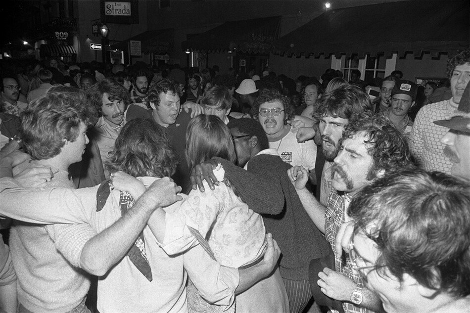 This grayscale photograph captures a crowded street scene at night with a group of people clustered together with their arms around each other's shoulders. The people, mostly male, exhibit a range of facial expressions, and many of them have their mouths open. They wear casual clothing typical of a social gathering, such as shirts and sweaters. Surrounding the central group, a dense crowd fills the background under the bright streetlights, some facing us and others looking away. Behind them are the facades of buildings showing store signage, including one reading "La Strada".