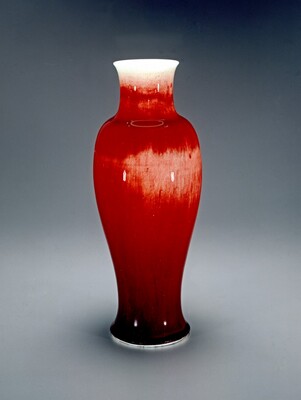 This large ceramic vase has a deep red glaze that fades to white at the top rim. The vase has a smooth, slightly elongated shape with a narrow neck and a round base. Around the middle section of the vase there is a lighter red section, and towards the bottom the red transitions into a dark, almost black shade.