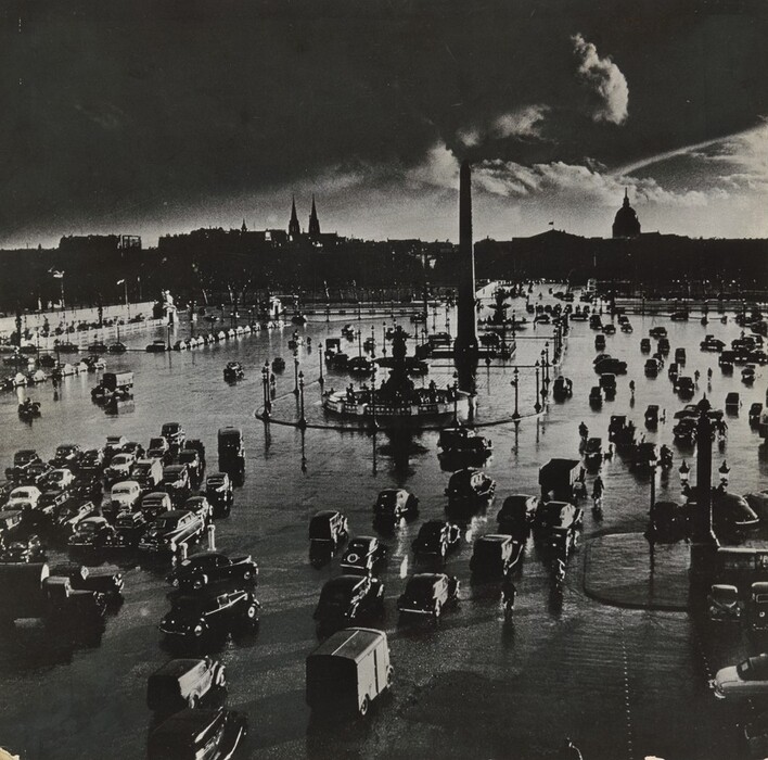 This is a photograph of a city square with cars, an obelisk, a fountain, and street lamps. A building with a dome and spires is visible against a cloudy sky. The image is in black and white.