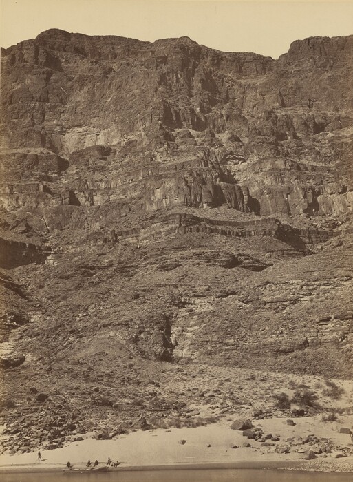 The image shows a cliff face viewed from a riverbank. The cliff rises steeply from the water, with a group of people and a small boat nearby. The rock surface is textured and layered, showing striations. The color palette includes earthy tones of brown and tan, with light and shadow enhancing the rugged appearance of the cliff.