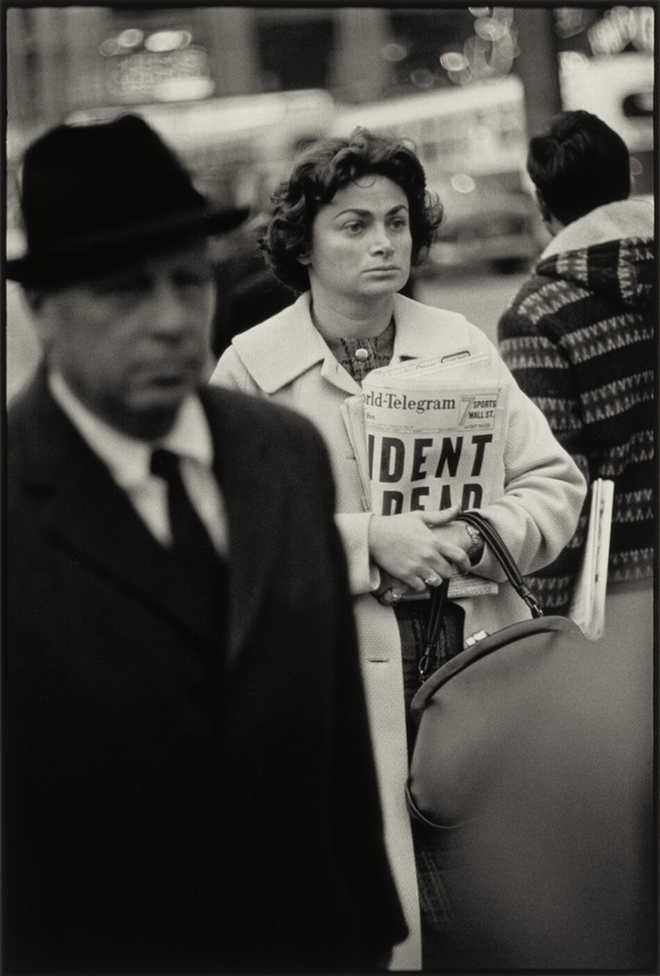 The image shows a woman positioned slightly off-center, facing forward. She has dark, curly hair, defined eyebrows, a straight nose, and full lips. The woman is wearing a light-colored coat over a patterned dress or blouse, and she is holding a rolled newspaper with the headline visible. To her right, another individual's shoulder clad in a patterned jacket is partially visible. The background is an urban setting with blurred lights and shapes, suggesting a bustling city atmosphere.