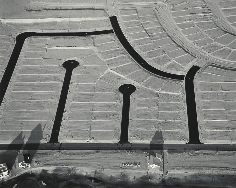 We look down onto plots of land divided and subdivided around streets and cul-de-sacs in this horizontal black and white photograph. A main access street runs across the bottom edge of the photograph. Three streets connect to make a rough square off the main road, and two round-ended cul-de-sacs reach up in between.