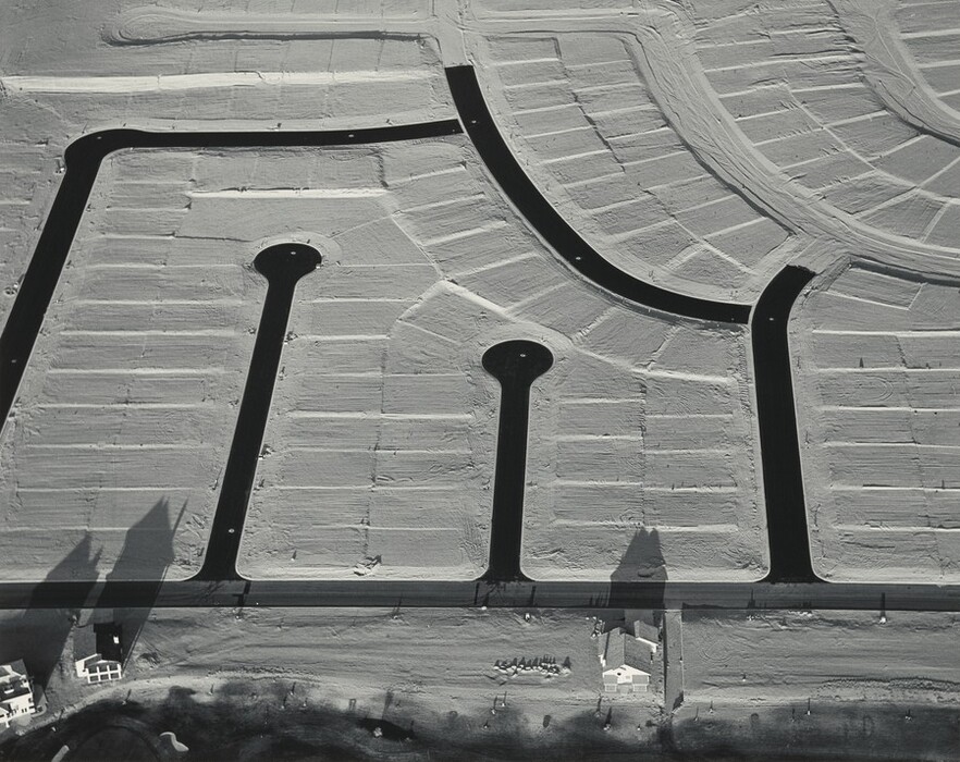 We look down onto plots of land divided and subdivided around streets and cul-de-sacs in this horizontal black and white photograph. A main access street runs across the bottom edge of the photograph. Three streets connect to make a rough square off the main road, and two round-ended cul-de-sacs reach up in between.