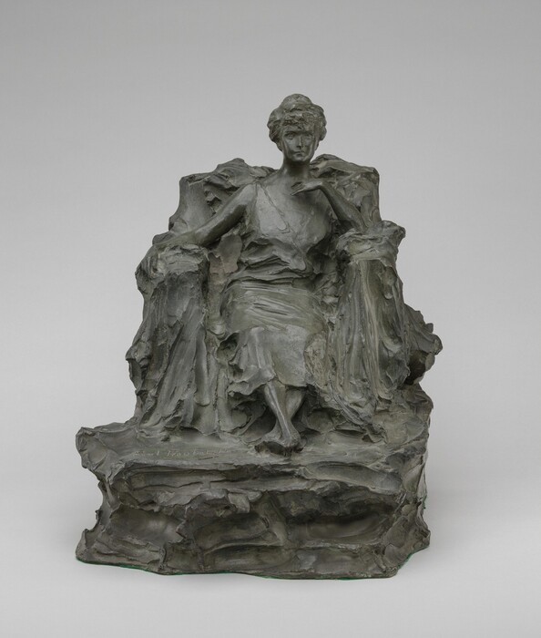 The sculpture depicts a seated figure of a woman draped in a flowing, classical-style garment. She is positioned slightly turned, with one arm resting on the arm of her seat and the other hand raised to her chest. The sculpture is composed of a bronze material. The woman's features are delicately rendered, emphasizing the contours of her face and the texture of her robe. The figure appears to be seated on a rugged, rock-like base. Overall, the sculpture portrays the woman in a poised yet relaxed demeanor, capturing a moment of serene contemplation.